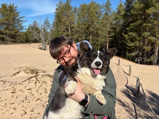 A black and white corgi cardigan being held be her dog dad (me!). She looks sandy and there is a sandy dune forest in the background.