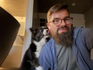 In the somewhat unsharp photo, there is a a beared and bespectacled man (me!) sitting on the floor and a dog (napu!). The dog has climbed up the arm of the man and is licking his ear